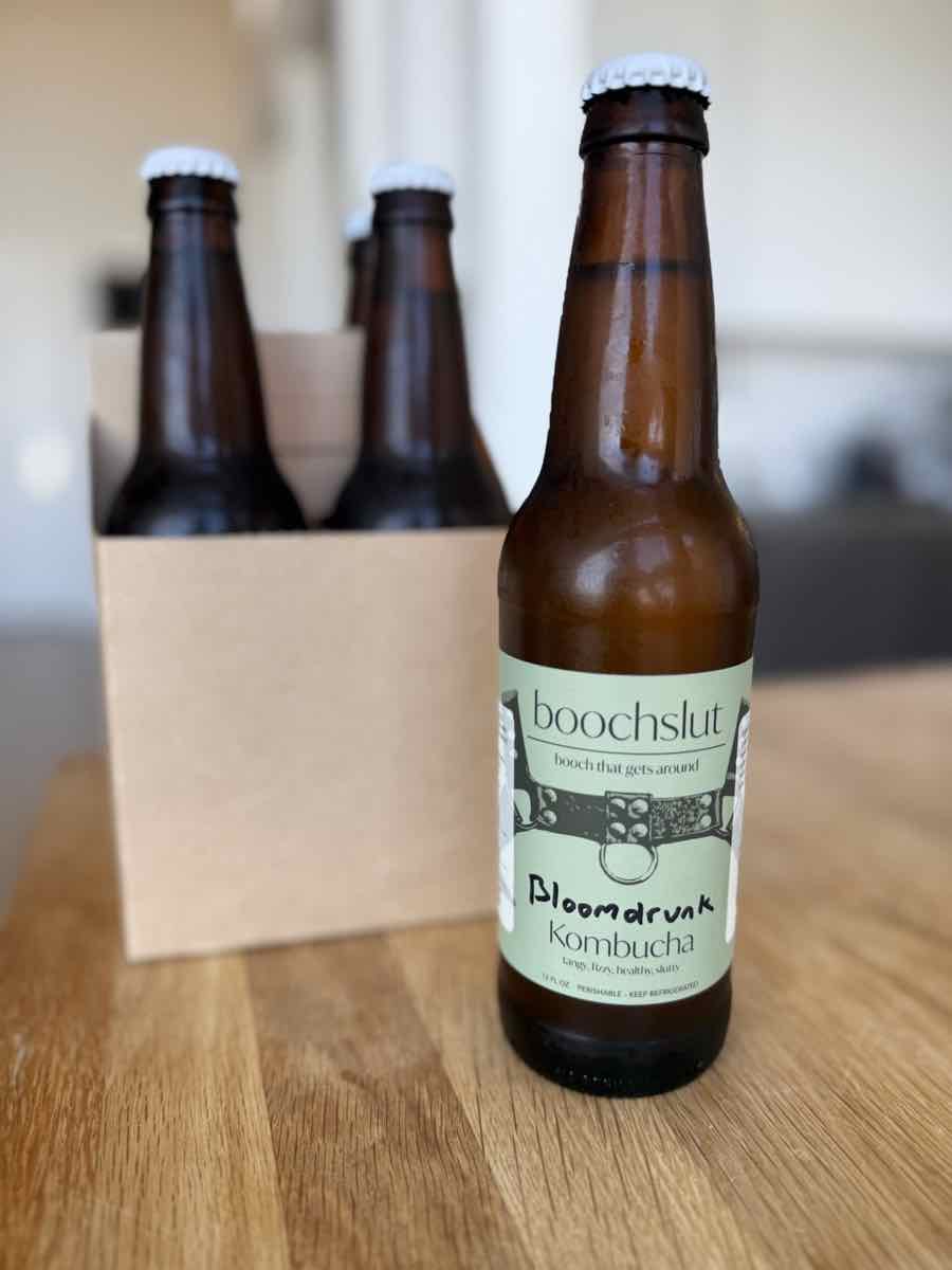 A bottle of boochslut Bloomdrunk kombucha in front of a four-pack carrier